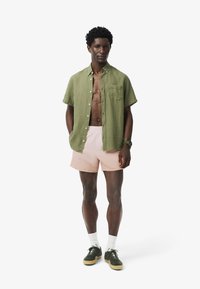 Camicia verde a maniche corte con bottoni, pantaloncini da bagno rosa, calzini bianchi e scarpe da ginnastica verde scuro. Tessuto liscio e vestibilità rilassata. Nessun motivo.