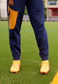 Pantaloni tuta sportiva della marina con accenti laterali arancioni e tasche con zip, abbinati a scarpe da calcio arancioni brillanti con design texturizzati.