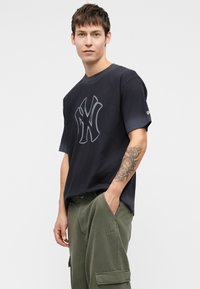 T-shirt de algodão preto com um grande logotipo cinza de NY no peito. Mangas curtas e decote redondo. Combinado com calças cargo verde-oliva.