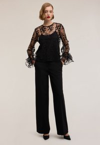 Top nero trasparente con dettagli floreali ricamati, indossato sopra una canottiera nera solida, abbinato a pantaloni neri a gamba larga.