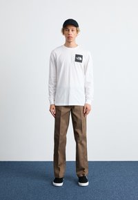 White long-sleeve shirt with black logo on chest, brown pants, and black cap. Black shoes with white soles. Simple, casual outfit.