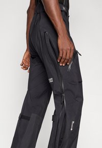 Czarne wodoodporne spodnie wykonane z lekkiego materiału, z zamkami błyskawicznymi wentylacyjnymi, akcentami z rzepem oraz detalami logo na udzie.