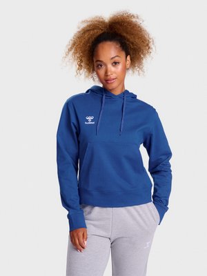 Vrouw met krullend haar die een blauwe hummel hoodie en lichtgrijze joggingbroek draagt, poseert met één hand in haar zak tegen een witte achtergrond.