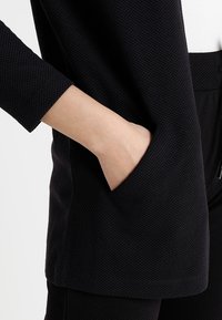 Hand gestoken in de zak van een gestructureerde zwarte jas, met bijpassende zwarte broek zichtbaar.