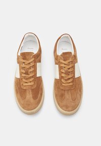 Sneakers in suede marrone con una striscia in pelle bianca, punta rotonda e suola in gomma beige. Presentano lacci piatti e dettagli cuciti.