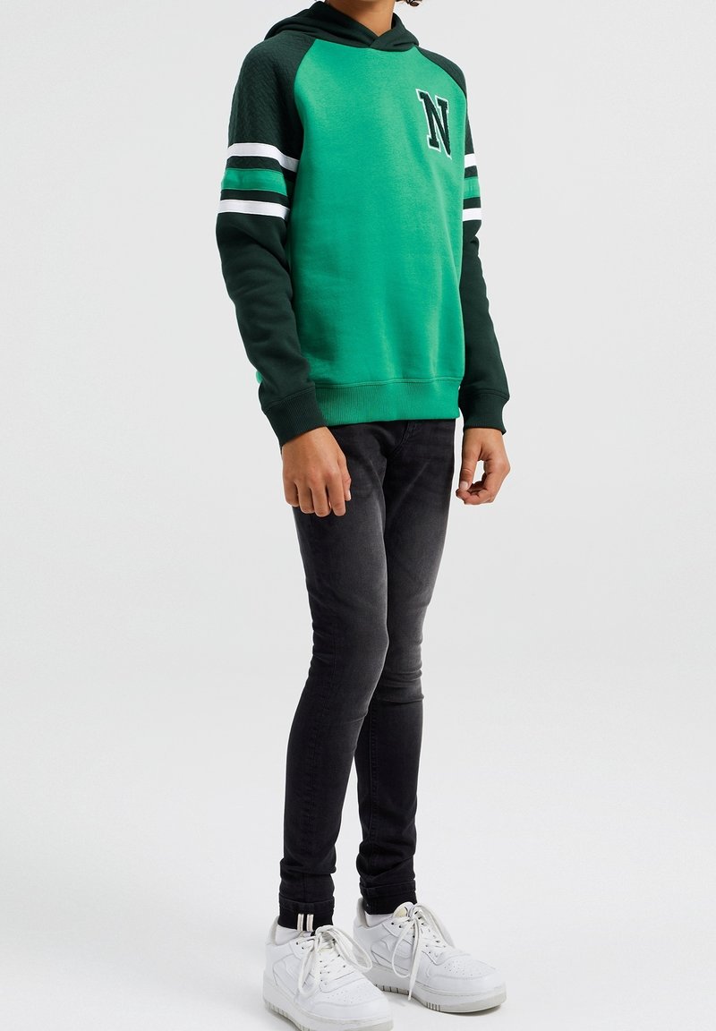 Person som bär en grön och svart hoodie med "N" på bröstet, svarta slim fit jeans och vita sneakers som står mot en enkel bakgrund.