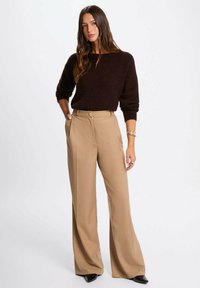 Maglione marrone a maglia con vestibilità ampia, abbinato a pantaloni crema a vita alta. I pantaloni presentano un dettaglio con bottone e un design a gamba larga. Scarpe nere.