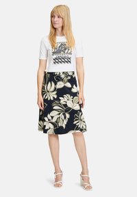 Weißes Kurzarm-T-Shirt mit "HAPPY" Grafik, kombiniert mit einem navyblauen, blumigen Midirock mit großen grünen und weißen Blumen. Weiße Absatzsandalen.