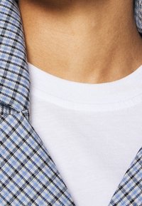 Blå och svart rutmönstrad jacka med krage över en vit t-shirt. Fokusera på textur och mönsterdetaljer, med en slät halsringning synlig.