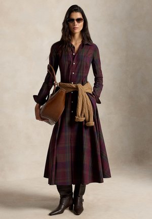 Femme portant une longue robe à carreaux boutonnée, des bottes marron, des lunettes de soleil, un pull beige noué à la taille, et tenant un grand sac à main en cuir marron.