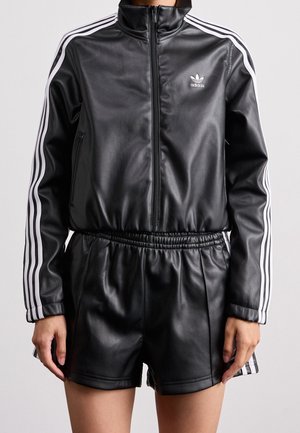 Osoba nosząca czarną, skórzaną kurtkę Adidas z białymi paskami na rękawach oraz dopasowane czarne, skórzane szorty na jednolitym tle.