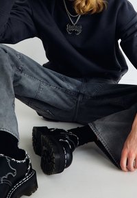Felpa nera a maniche lunghe, jeans grigi a gamba larga con superficie texturizzata e scarpe chunky nere con dettagli di cucitura bianca.