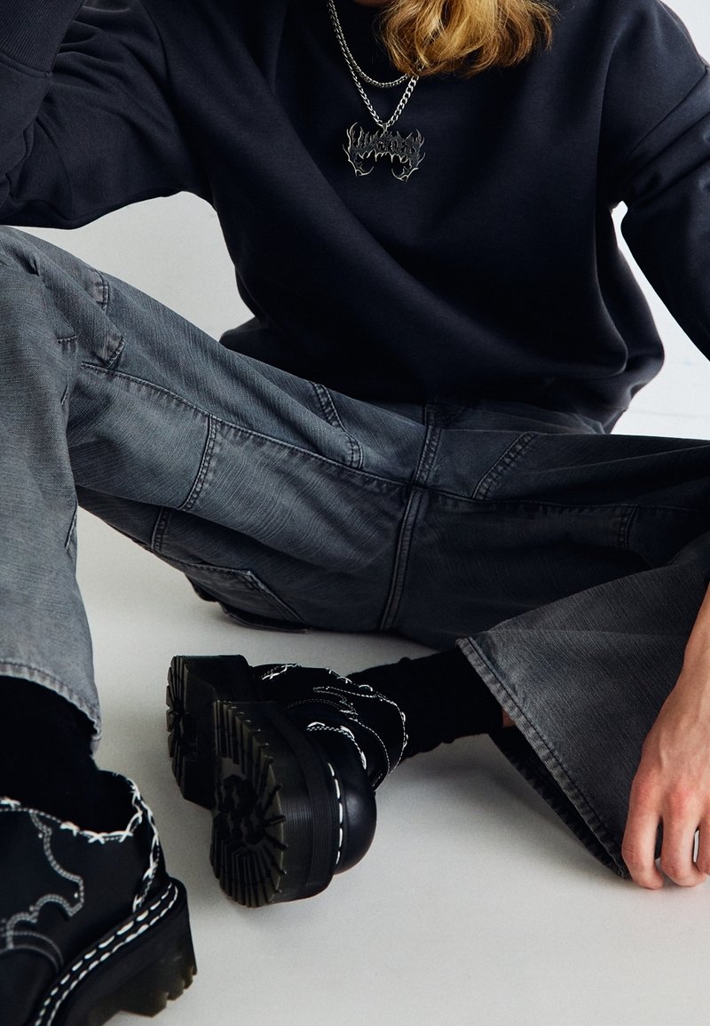 Felpa nera a maniche lunghe, jeans grigi a gamba larga con superficie texturizzata e scarpe chunky nere con dettagli di cucitura bianca.