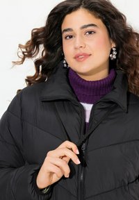 Svart puffjacka med kviltad struktur, hög krage och dragkedjestängning, bärs över en lila polo. Silverfärgade statementörhänge.