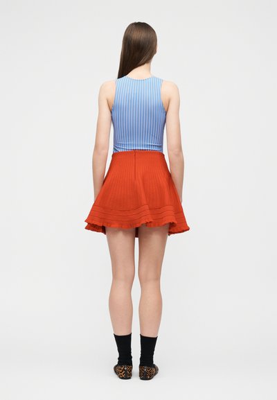 Femme debout de dos portant un haut sans manches à rayures bleues, une jupe plissée rouge, des chaussettes noires et des chaussures à imprimé léopard.