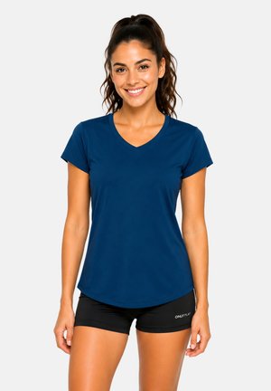 Lächelnde Frau mit dunklen Haaren im Pferdeschwanz, die ein blaues T-Shirt mit V-Ausschnitt und schwarze Shorts trägt, steht vor einem schlichten hellen Hintergrund.