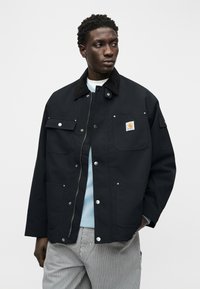 Άνδρας που φοράει μαύρο μπουφάν Carhartt με εμπρόσθιες τσέπες, ανοιχτόγαλάζιο πουκάμισο και ριγέ παντελόνι, κοιτώντας προς τα πλάγια, με μονόχρωμο φόντο.