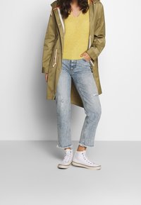 Imperméable vert olive avec capuche à cordon, pull en tricot jaune, jean déchiré bleu clair et baskets montantes blanches.