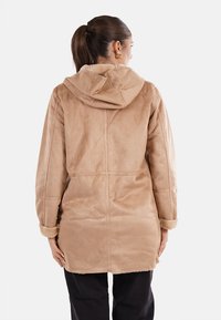Cappotto in pelliccia sintetica beige con cappuccio, caratterizzato da una texture morbida, maniche lunghe e una vestibilità leggermente ampia. Mostra la vista posteriore con cuciture verticali.