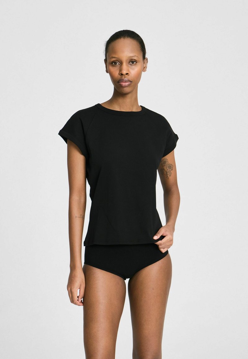 Moteris su trumpais juodais plaukais, dėvinti laisvą juodą marškinėlį ir juodus apatinukus, stovinti priešais paprastą šviesiai pilką foną.