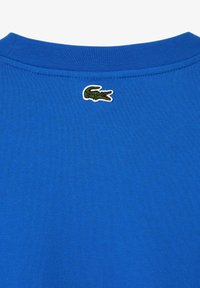 Camiseta de algodón azul con cuello redondo acanalado y logo de cocodrilo verde bordado en la parte posterior del cuello. Textura suave.