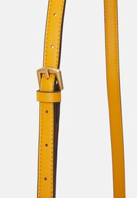 Gelber Lederarmband mit schwarzem Rand, ausgestattet mit einer goldenen Schnalle und verstellbaren Rillen für die Größe. Glatte Textur und sichtbare Nähdetails.