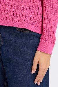 Pull en tricot fuchsia avec un motif texturé et un ourlet côtelé, associé à un jean en denim bleu foncé. Main visible reposant sur le jean.