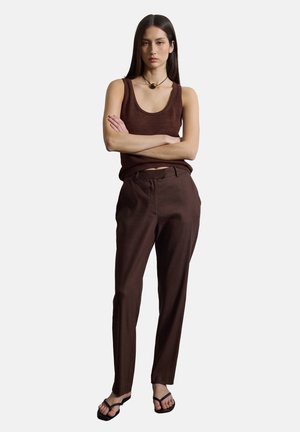 Frau steht mit verschränkten Armen, trägt ein ärmelloses braunes Top, braune Hose, schwarze Sandalen und eine runde Anhänger-Halskette.