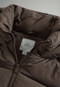 Brun pufferjacka med hög krage, med en framdragkedja och texturerat tyg. Etiketten visar "THE SET", storlek M, och skötselråd.