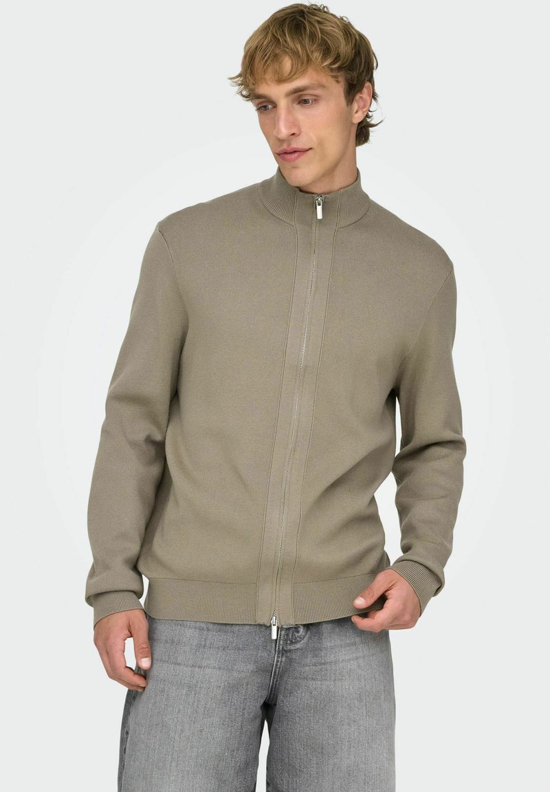 Maglione beige chiaro con zip, collo alto, polsini e orlo a coste, e tessitura a maglia fine, abbinato a jeans grigio chiaro.