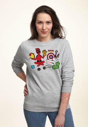 Kvinde iført grå sweatshirt med tegneserie-Deadpool, der holder Iron Man-maske og Infinity Gauntlet omgivet af Marvel-superheltegenstande og logo.