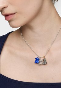 Srebrny łańcuszek z kulkami, na którym znajdują się trzy zawieszki: niebieski miś, teksturowana srebrna figura oraz ciemny miś z ozdobami.