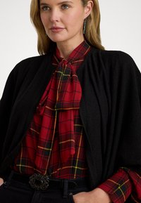 Cardigan negru drapat peste o bluză cu carouri roșii, având accente galbene și negre, cu un gât legat și o cataramă decorativă neagră.