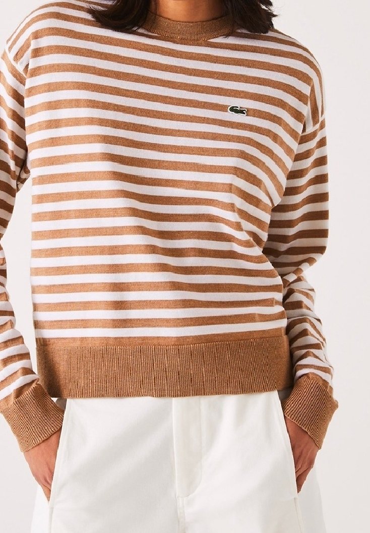 Personen bär en brun och vit randig tröja med ribbad mudd och nederkant, tillsammans med vita byxor, händerna i fickorna.