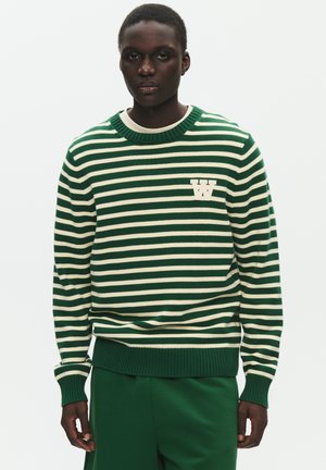 Person iført en grøn og hvid vandretstribet sweater med et "W"-logo på brystet, kombineret med ensfarvede grønne bukser.