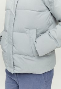 Personne portant une veste matelassée gris clair avec la main dans la poche latérale et un pantalon en velours côtelé bleu visible de la taille à la cuisse.