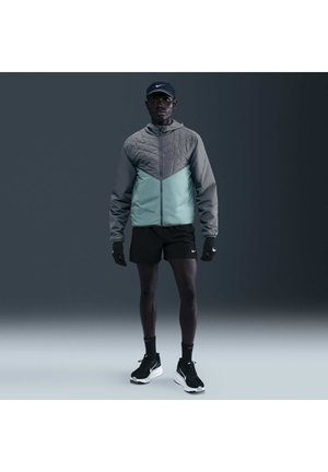 Mężczyzna ubrany w szarą i miętowo zieloną kurtkę, czarne spodenki, czarne rękawiczki, czarną czapkę Nike, skarpetki i czarne buty do biegania stojący na szarym tle.