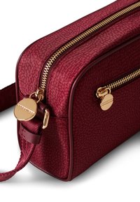 Burgundy rectangular bag made of textured fabric, featuring a double zipper and a gold-tone hardware detail on the front. Adjustable strap.