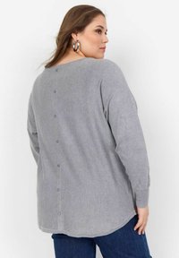 Grauer, langärmeliger Strickpullover mit abgerundetem Saum und Knopfdetail am Rücken. Weicher Textur, lockere Passform, getragen mit dunkelblauen Jeans.