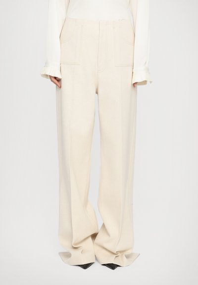 Pantalons beige à jambes larges avec deux poches frontales, tissu texturé et détails de couture subtils, associés à un haut à manches longues blanc.