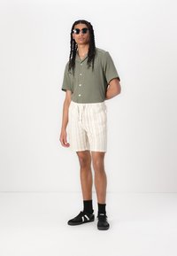Grøn ribbet kortærmet skjorte, beige stribede shorts og sorte sneakers med hvide accenter. Solbriller fuldender outfittet.