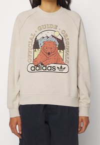 Ljus beige sweatshirt med rund halsringning, med en grafisk design av en björn, berg och träd. Adidas-logotyp längst ner. Mjuk struktur.