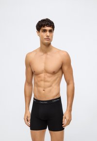 Een naakte man met kort, krullend haar die zwarte Calvin Klein boxer shorts draagt, staand tegen een effen lichtgrijze achtergrond.