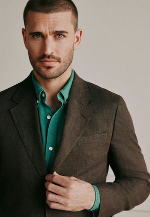 Muž s krátkymi tmavými vlasmi a bradou si upravuje hnedý blejzer cez zelenú košeľu na gombíky, pozerá sa priamo pred seba s neutrálnym výrazom.