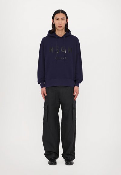 Felpa con cappuccio blu navy con logo frontale, abbinata a pantaloni cargo neri con tasche laterali e vestibilità comoda, indossata con scarpe nere.