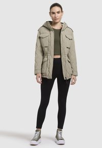 Khaki gewatteerde pufferjack met capuchon, trekkoord in de taille en vier voorzakken, gecombineerd met zwarte leggings en groene hoge sneakers.