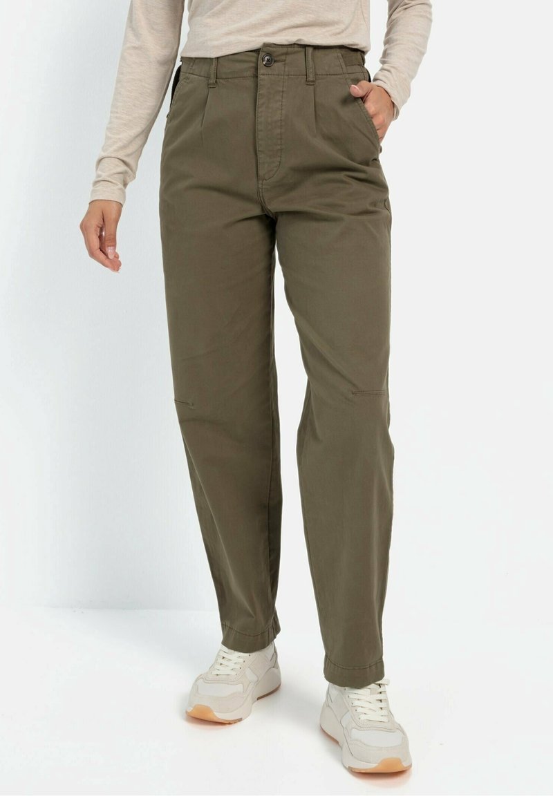 Personne portant un pantalon taille haute vert olive, un haut beige à manches longues et des baskets blanches, debout avec une main dans la poche.