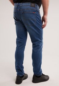 Mees, kes kannab siniseid kitsaid teksaseid ja musti tenniseid, seisab heleda tavalise tausta ees, seljataga vaade.