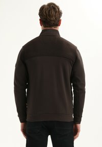 Donkerbruin sweatshirt met ribbelstructuur over de bovenrug. Voorzien van een rechtopstaande kraag en smalle manchetten, gemaakt van een zachte, gladde stof.