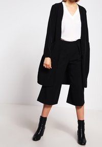 Cardigan lungo nero su una maglietta bianca con scollo a V, abbinato a culotte nere a gamba larga e stivali alla caviglia. Tessuto liscio, design minimalista.